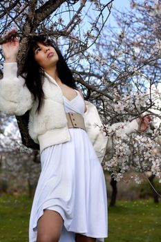 View of a beautiful girl on a white dress on a green grass field next to a almond tree