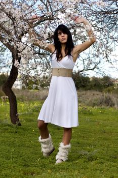 View of a beautiful girl on a white dress on a green grass field next to a almond tree