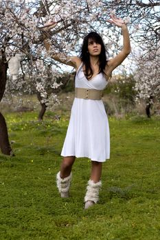 View of a beautiful girl on a white dress on a green grass field next to a almond tree