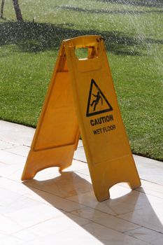 Yellow cleaning sign, caution wet floor, sprinklers on in the background