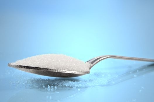 Close and low level capturing a tea spoon with sugar granules against a blue background.