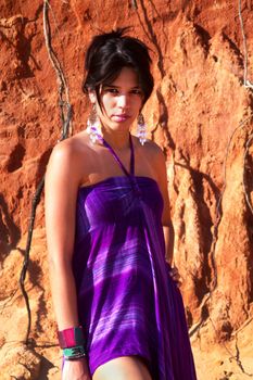 View of a beautiful young girl posing with a purple dress next to a cliff on the beach. 