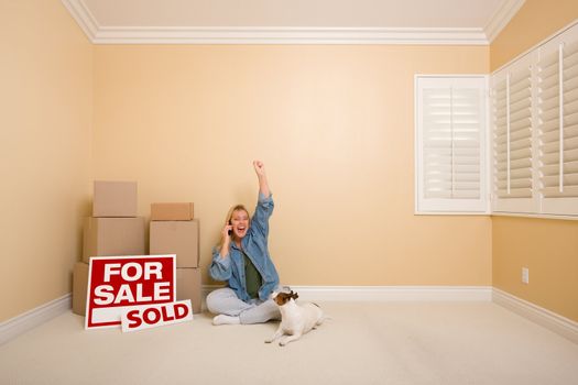 Pretty Woman Sitting on Floor Using Phone Celebrating Next to Moving Boxes, Sold Real Estate Signs and Dog in Empty Room.