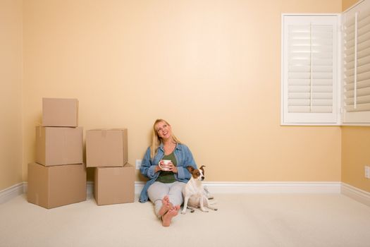 Pretty Woman Sitting on Floor with Cup Next to Moving Boxes and Dog in Empty Room.