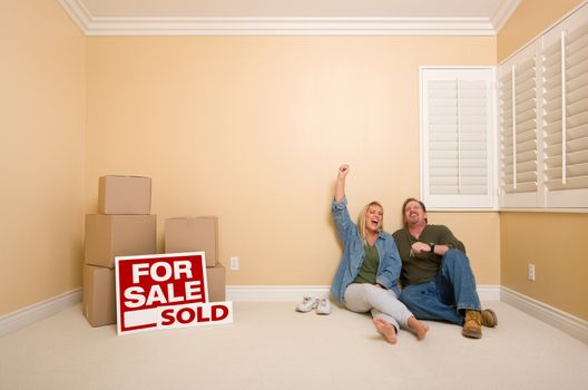 Excited Couple Relaxing on Floor Near Boxes and Sold Real Estate Signs in Empty Room.