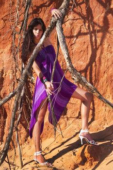 View of a beautiful young girl posing with a purple dress next to a cliff on the beach. 