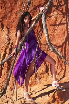 View of a beautiful young girl posing with a purple dress next to a cliff on the beach. 