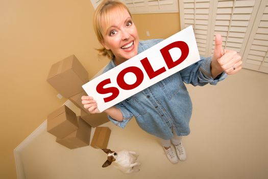 Excited Woman with Thumbs Up and Doggy Holding Sold Real Estate Sign Near Moving Boxes in Empty Room Taken with Extreme Wide Angle Lens.
