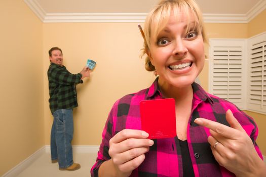 Fun Happy Couple Comparing Paint Colors in Empty Room - Woman Large, in Front, Man Smaller, Behind.