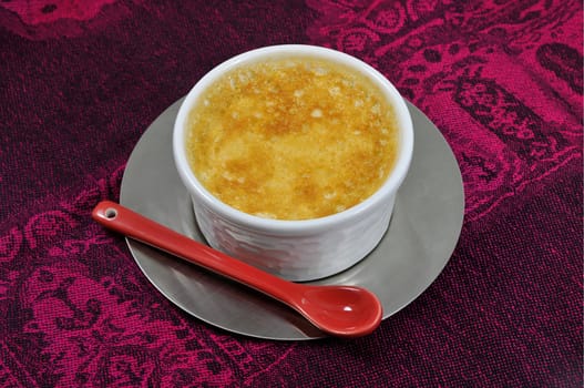 Custard with a red ceramic spoon and an iron steel saucer on a magenta tablecloth