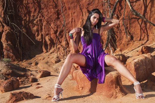 View of a beautiful young girl posing with a purple dress next to a cliff on the beach. 