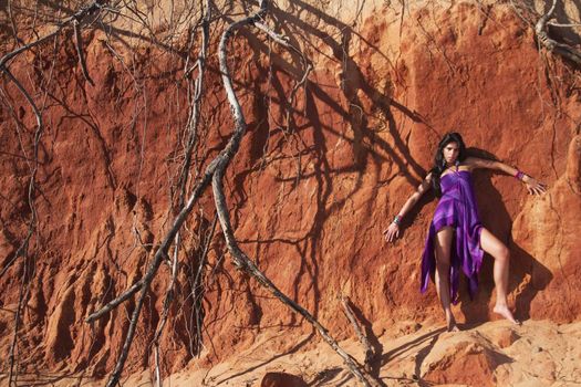 View of a beautiful young girl posing with a purple dress next to a cliff on the beach. 