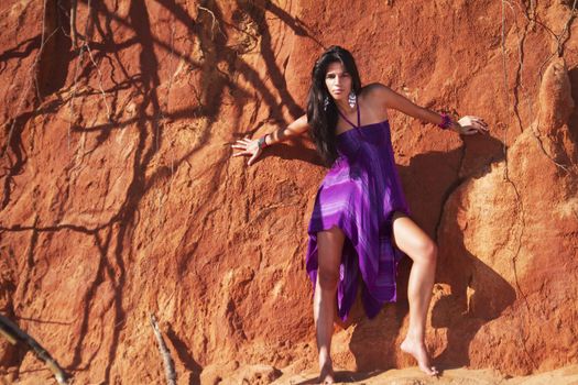 View of a beautiful young girl posing with a purple dress next to a cliff on the beach. 