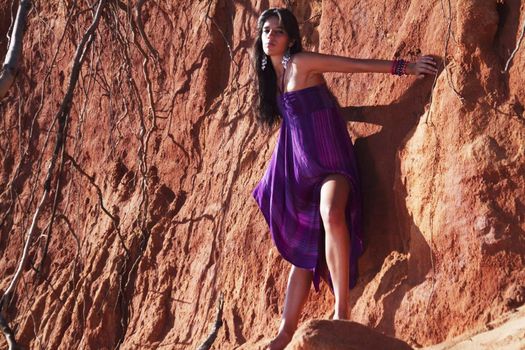 View of a beautiful young girl posing with a purple dress next to a cliff on the beach. 