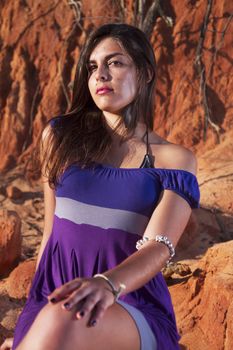 View of a beautiful young girl with a purple dress standing on a red cliff on the beach.