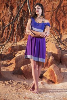 View of a beautiful young girl with a purple dress standing on a red cliff on the beach.