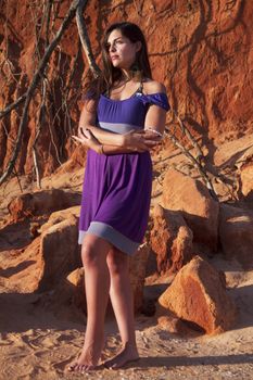 View of a beautiful young girl with a purple dress standing on a red cliff on the beach.