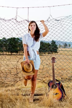 View of a beautiful young country girl with a guitar on the fence of the grassland. 