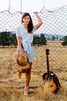 View of a beautiful young country girl with a guitar on the fence of the grassland. 