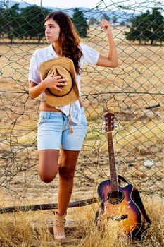 View of a beautiful young country girl with a guitar on the fence of the grassland. 