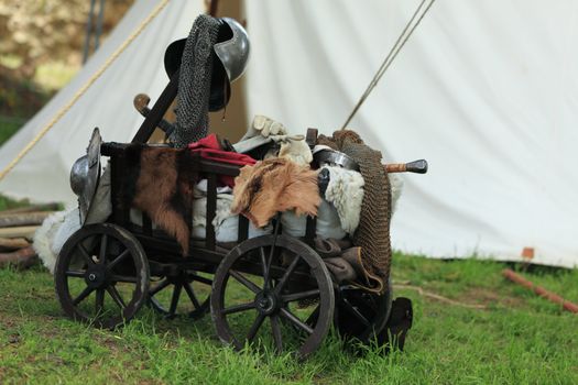 A wooden old cart full of medieval military stuff near a traditional campaign tent.