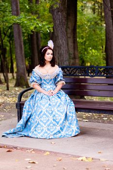 A portrait of lady in a blue baroque dress sitting on bench
