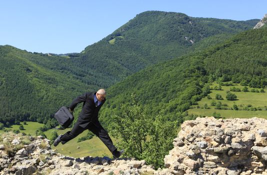 Businessman with a laptop bag running outdoors on a rocky ruined stronghold wall in a mountaineous area.The image can suggest the idea of a thief stealing important data or a man protecting data in case of a disaster.