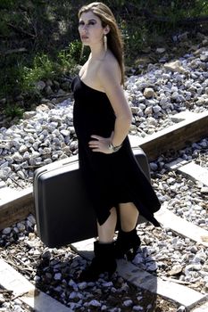 View of a beautiful woman with black dress with a travel case on a train track.