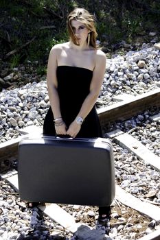 View of a beautiful woman with black dress with a travel case on a train track.