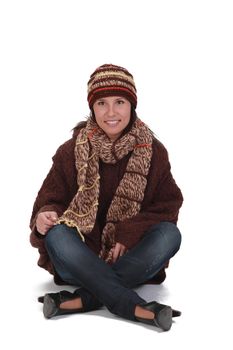 Happy young woman in winter clothes sitting crosslegged against a white background.