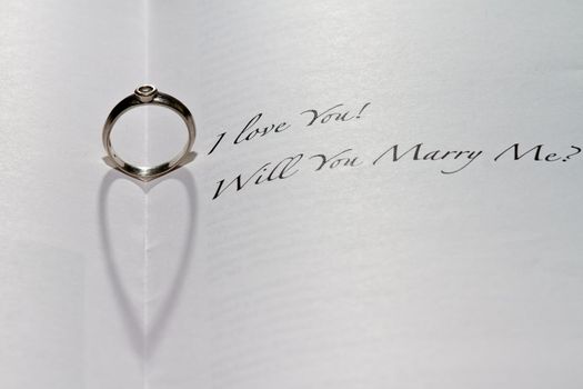 Ring casting heart shadow in book with the sentence "I Love You! Will You Marry Me?"