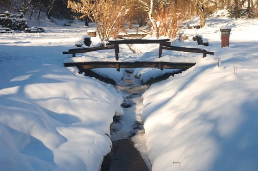 Wooden bridge over small frozen brook with snow and ice