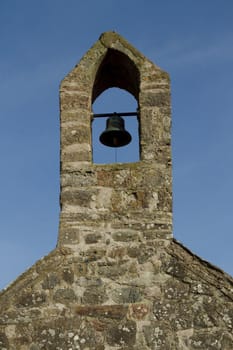 Traditional stone wall and tower houses a bronze bell with bar and rope against a blue sky.