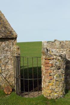 Metal swing gate with chain and weight between walls leading to a green grass field and light blue sky