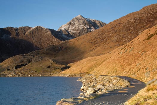 A footpath curves around a mountain lake with steep grass banks and rocky mountain cliffs in the distance.