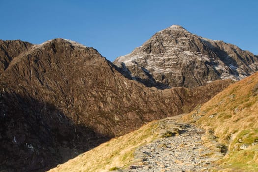 A footpath leads up past a grassy bank towards large mountainous cliffs with ridgelines and peaks.