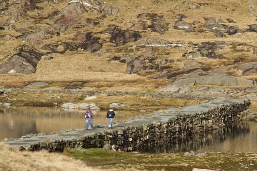 Two walkers cross a stone built causeway, with a footpath, between two bodies of water.