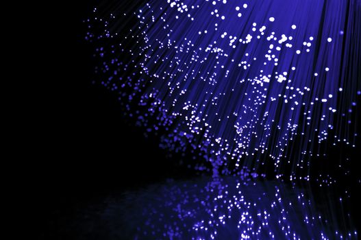 Close up on the ends of many illuminated fibre optic strands against black and reflecting into foreground.