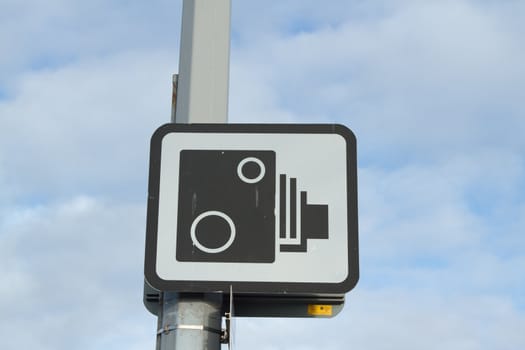 Symbol of a spped camera in black, on white, on a square sign attached to a post against a cloudy sky.