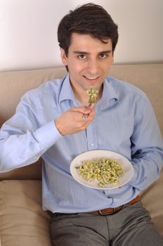 attractive young man sitting on the couch eating lunch (pasta with chicken)
