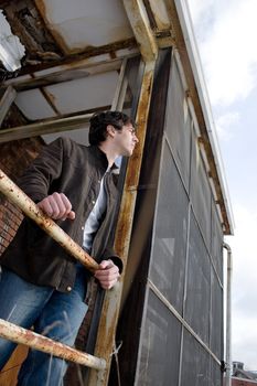 A young brunette man posing outside a worn and abandoned building.