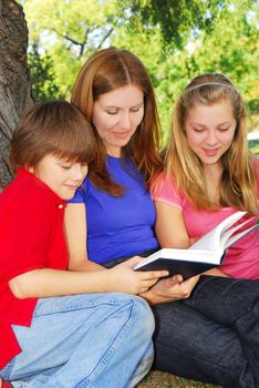 Family of mother and children reading a book under a tree in summer park