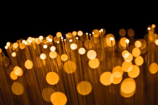 Close up on the ends of many illuminated golden fibre optic strands with many unfocussed bokeh effects against black background.