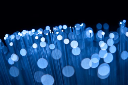 Close up on the ends of many illuminated blue fibre optic strands with many unfocussed bokeh effects against black background.