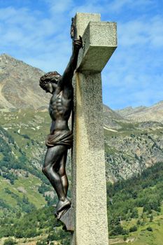 Side of a metallic sculpture of Jesus-Christ on a stone cross next to a mountain