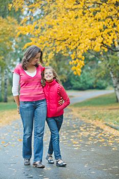 Cute girl with her mother walking in the autumn park. Rain, yellow leaves, tree.