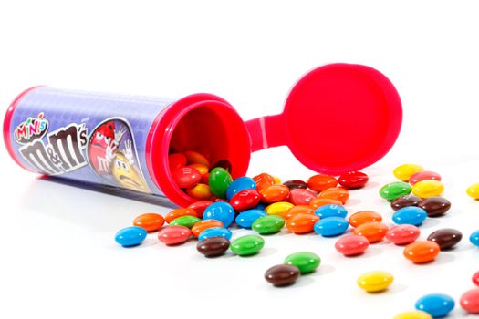 An open container of M&M Minis (chocolate button shaped candies) spilling out and scattered across a white backdrop.  Over 100 minis in each tube.  Made by Mars Inc.  Editorial Use Only