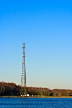 A communication tower at the edge of a lake with a clear blue sky as the background. Photo is in vertical or portrait format.