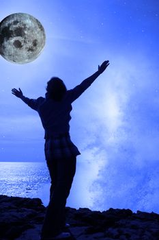 a lone woman raising her arms in awe at the powerful wave and full moon on the cliffs edge in county clare ireland