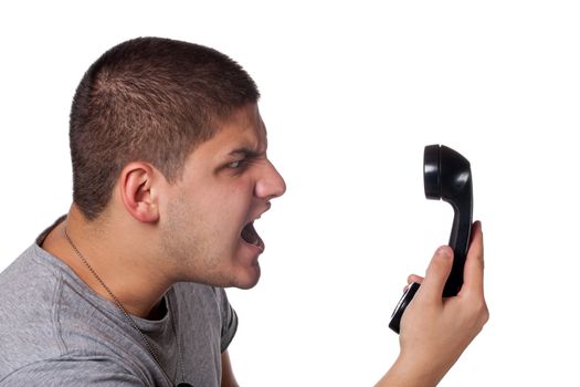 An angry and irritated young man yells into the telephone receiver over a white background.